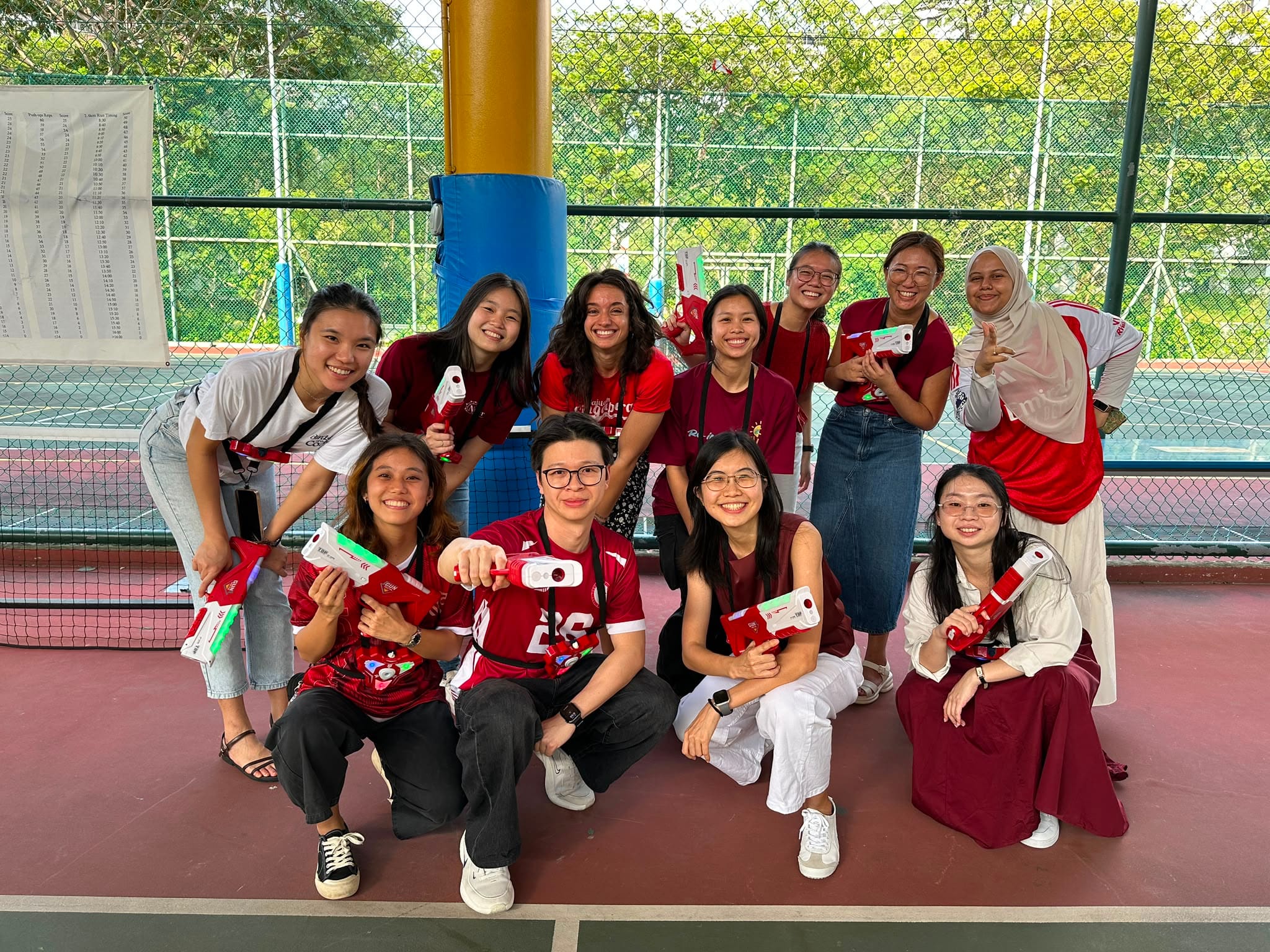 A group of smiling teachers pose together on an outdoor court, holding laser tag guns in red and white.